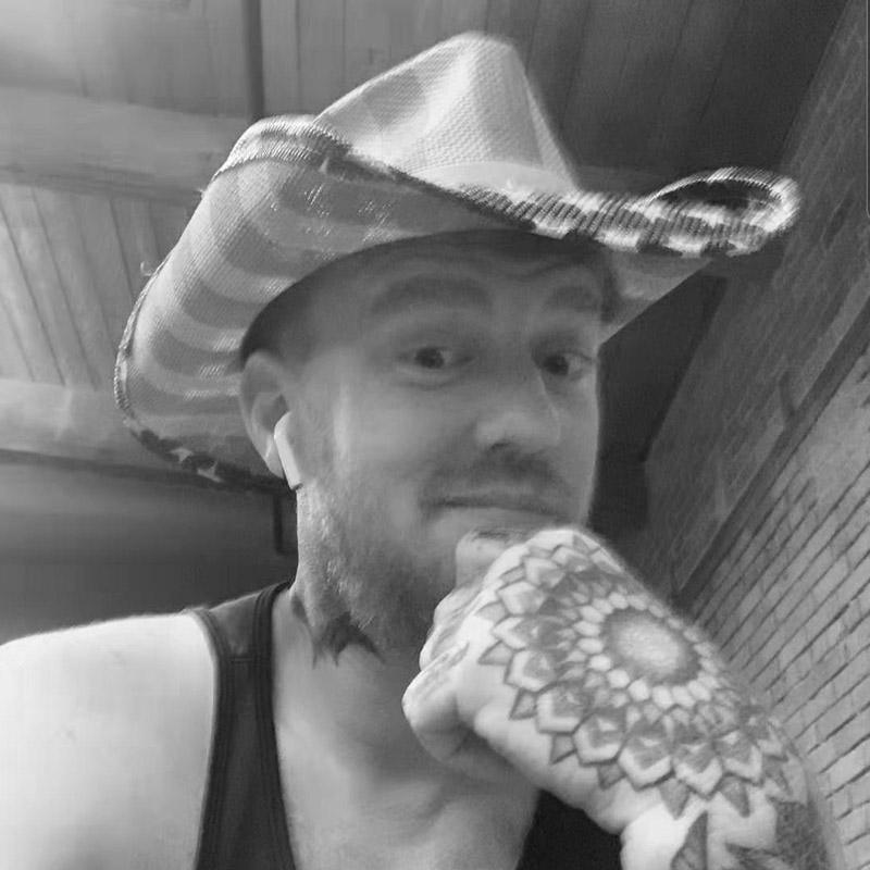 Tattooed man in cowboy hat with mandala hand tattoo, brick wall background, FYT Tattoo Supplies New York